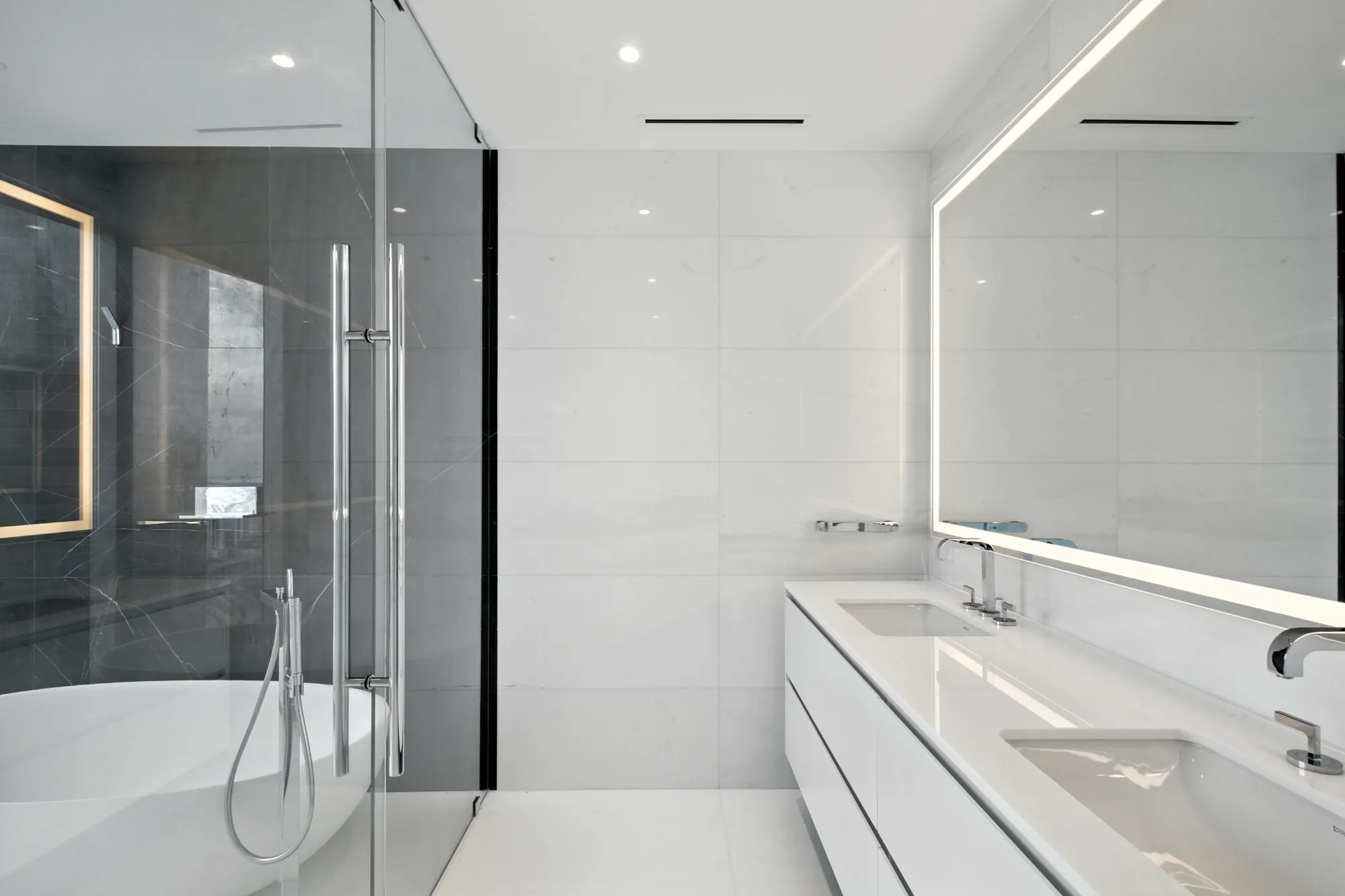Modern and chic luxury master bathroom at Aston Martin Residences. Double vanity on the right with enclosed bathtub and shower area on the left. Large mirror with light around the edges.
